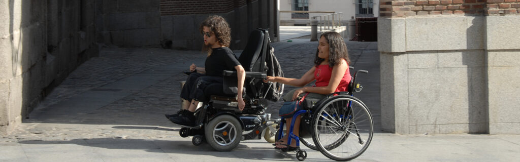 A young woman using a manual wheelchair hangs onto the back of a young man's power wheelchair as he pulls her along a street.