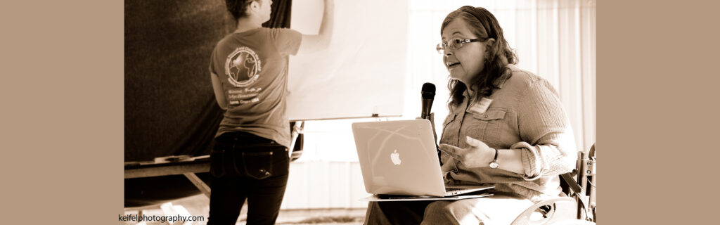 A woman in a wheelchair speaks into a microphone and holds a laptop in her lap. A woman writes on a flip chart in the background.