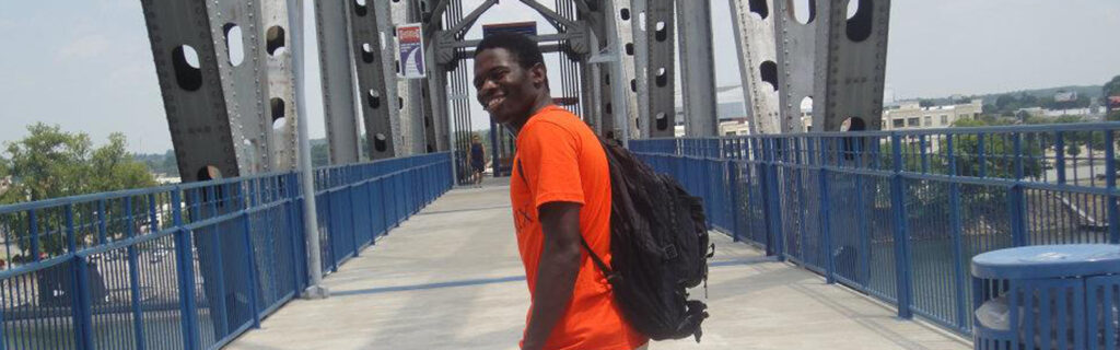 Samson, wearing a bright orange shirt, poses for a picture in front of a bridge.