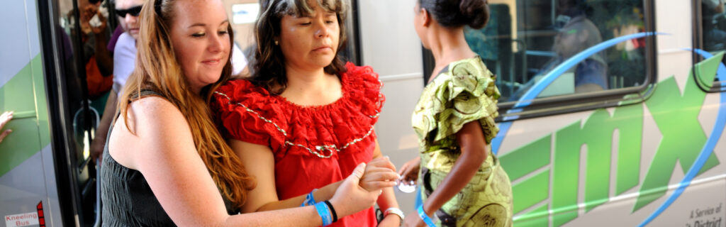 A young Latin American blind woman exits a public bus as a sighted American woman guides her.