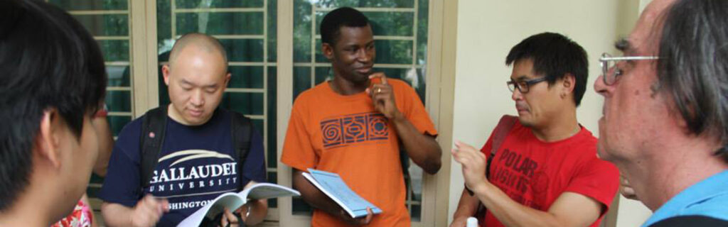 A group of male international college students communicate in sign language. One wears a Gallaudet t-shirt.