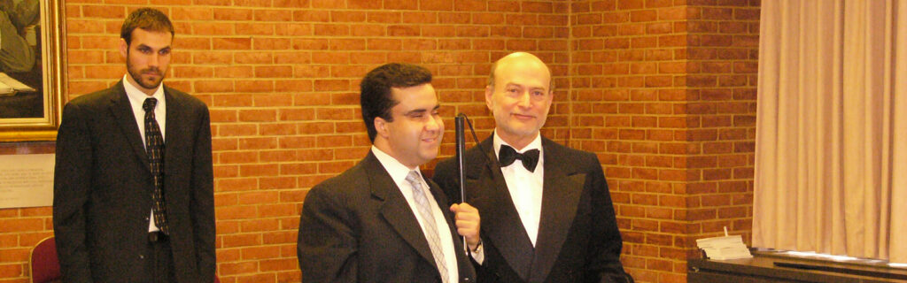 A smiling blind man in a suit holds a cane while greeting a man in a formal suit.