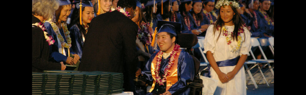 An international student from Asia in a power wheelchair wears a graduation cap and gown as he accepts his diploma.