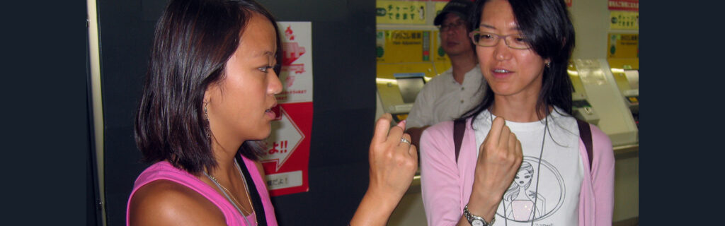 Two women are pictured. One of the women is showing the other how to form a particular sign.