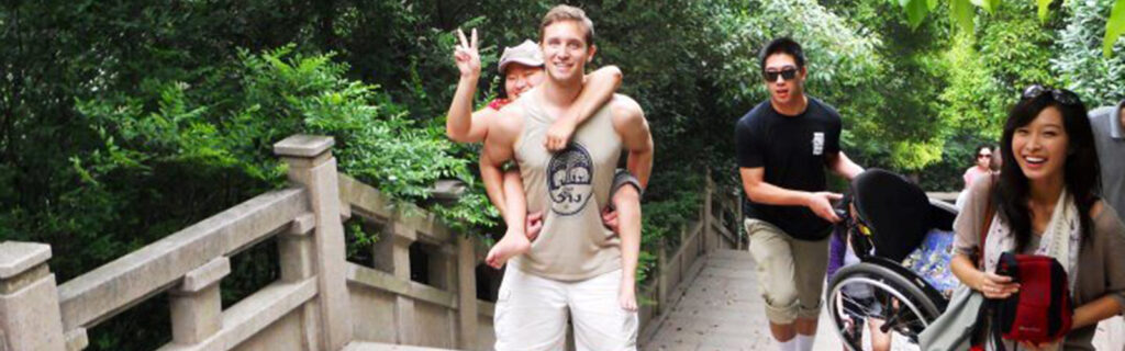 A woman is carried up stairs on someone's back, while others bring her wheelchair.