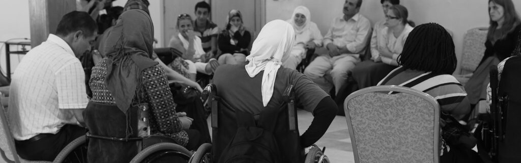 Black and white photo of people, including several women wheelchair users, sitting in a circle listening to a speaker.