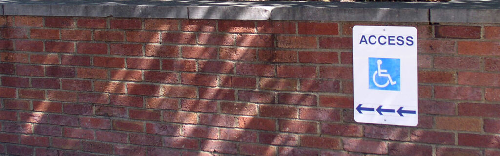 The sign with the symbol for wheelchair access is posted on a brick wall outside of a building.