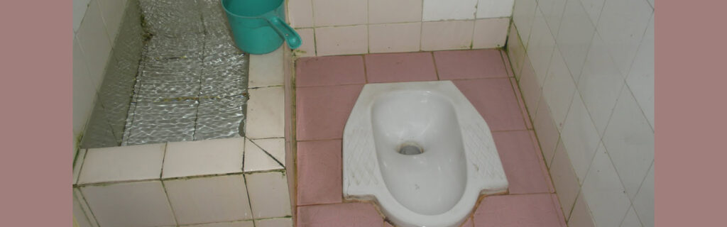 Toilet in the tiled floor with places to step while squating over the bowl, and a basin of water is next to it.
