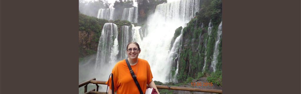 Emily smiling while standing in front of large waterfall holding her white cane.