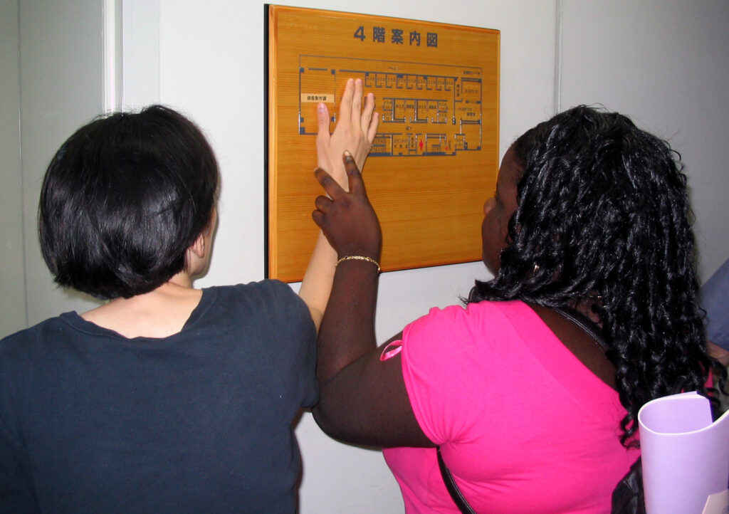 A woman is taking the hand of a blind woman to let her feel a tactile map mounted on the wall.