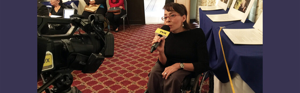 A women using a wheelchair speaks into a microphone in front of a TV camera.