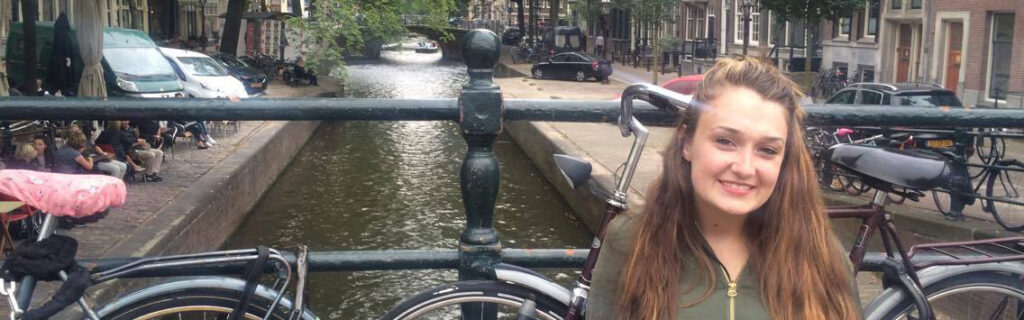 young woman sits on the bridge of a canal, with bicycle and canal visible behind her