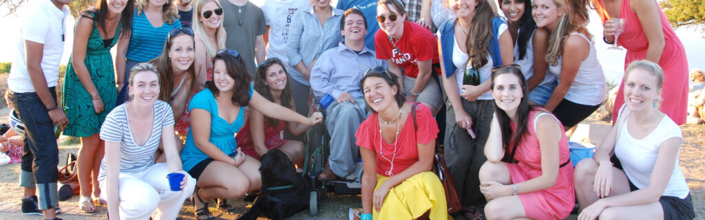 Crowd of over 20 young people, mostly non-disabled. Among them is a young man who uses a power wheelchair and his service dog.