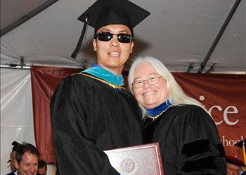 Chart is a middle-aged southeast Asian man who wears graduation regalia and dark sunglasses and an older woman with long white hair leans against him smiling.