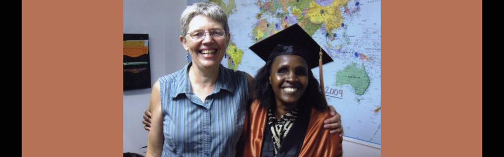 Flavia in graduation regalia smiles big standing next to her teacher with world map in the background.