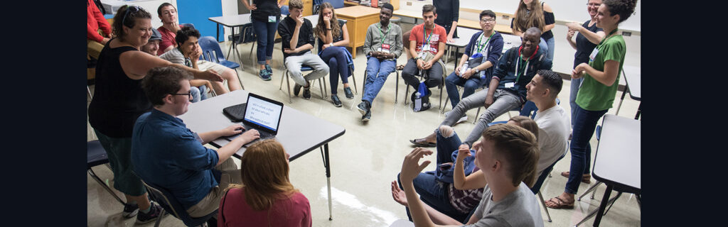 A large group of international students laughing as they sit in a circle in their chairs in a classroom. There is a man typing in front of the circle and there is a woman to the right using hand gestures to signal to one student, as he looks to her using sign language.