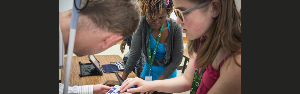 A man holding a white cane demonstrates how to use a refreshable braille display to two girls.