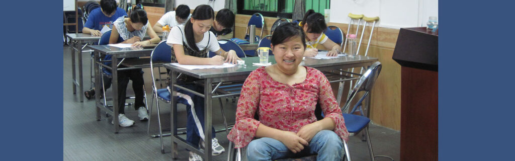 Ming sitting in wheelchair in front of classroom as students behind her have their heads down writing.