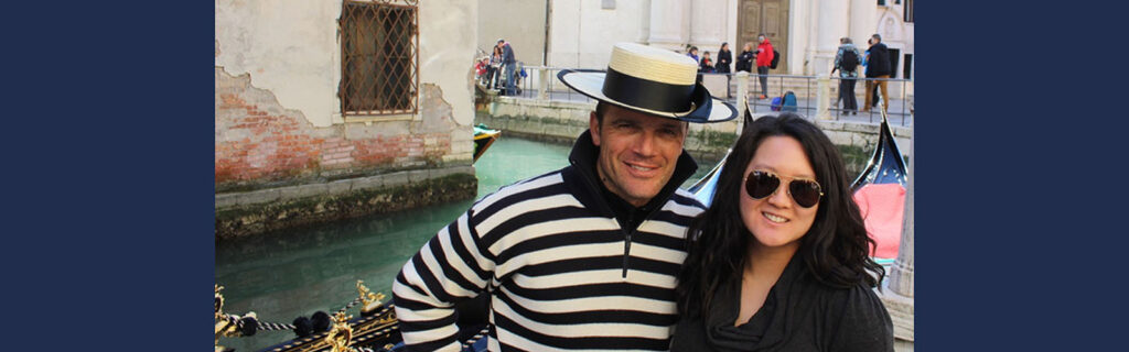 Sheila standing with a gondolier guide in front of canal in Venice with a gondolier in the background.