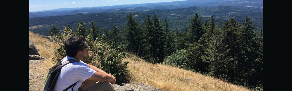 Cheng sitting on a mountain top overlooking view of trees and blue skies.