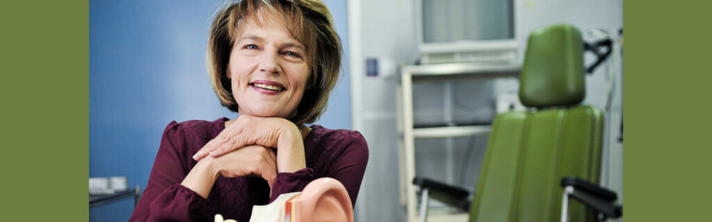 Magteld smiles and rests her chin on folded hands. Exam chair in background