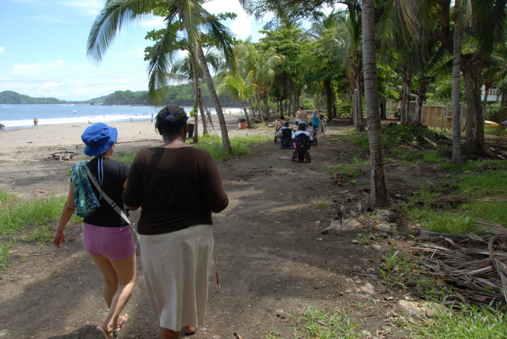 Two young women walk together along a jungle path parallel to a beach. One woman is carrying a white cane.