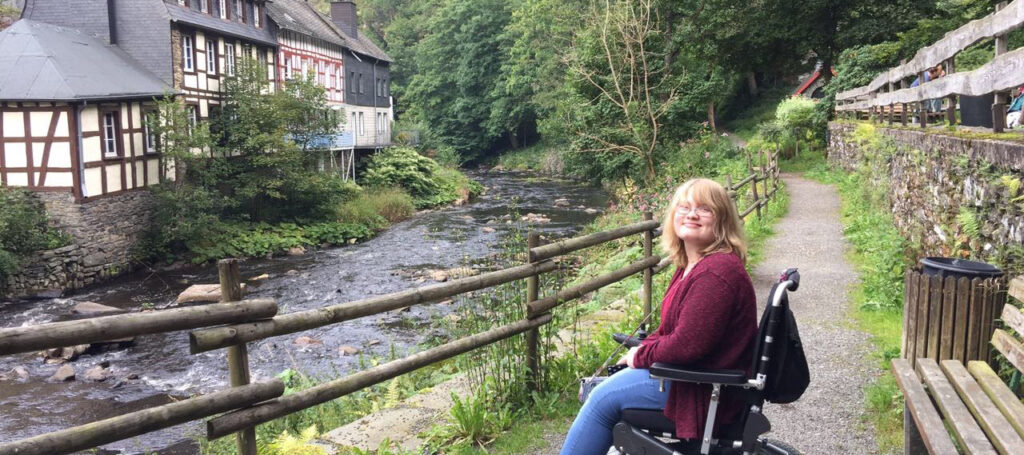 Brooklyn, seated in an electric wheelchair on a gravel path, faces a narrow river and German house