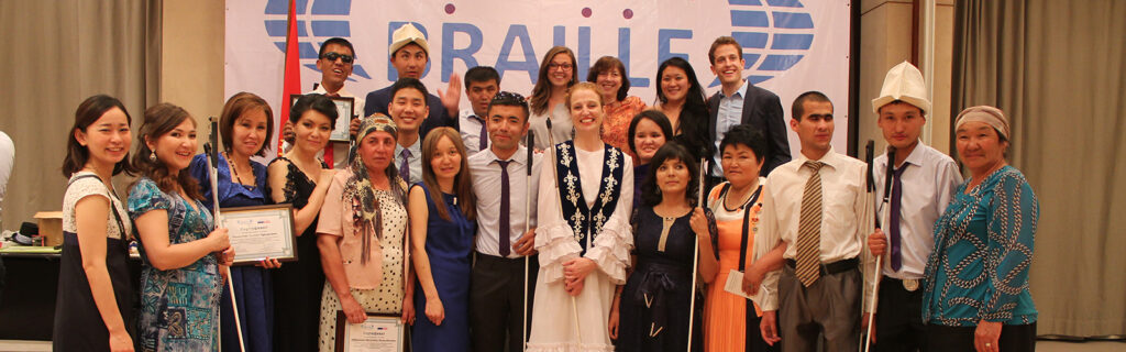 A white woman stands wearing a traditional Kyrgz dress, holding her white cane with a group of blind Kyrgz students in front of a banner that reads "Braille".