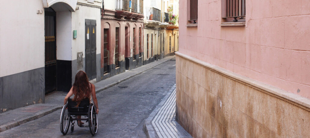 A young woman with long hair pushes herself in a manual wheelchair through a narrow alley in Spain painted in pastel colors