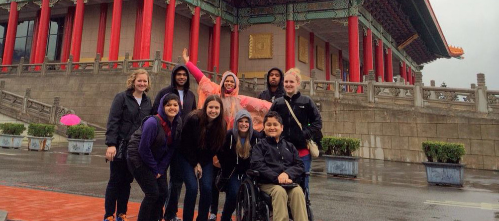A group of male and female students, including a man seated in a power wheelchair, gather in front of a Chinese monumen in the rain.