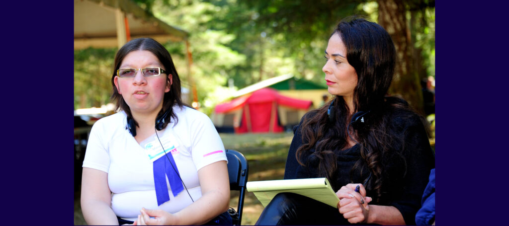 Two women sitting side by side in the outdoors with headphones around their neck, and one woman holding a notepad and pen.
