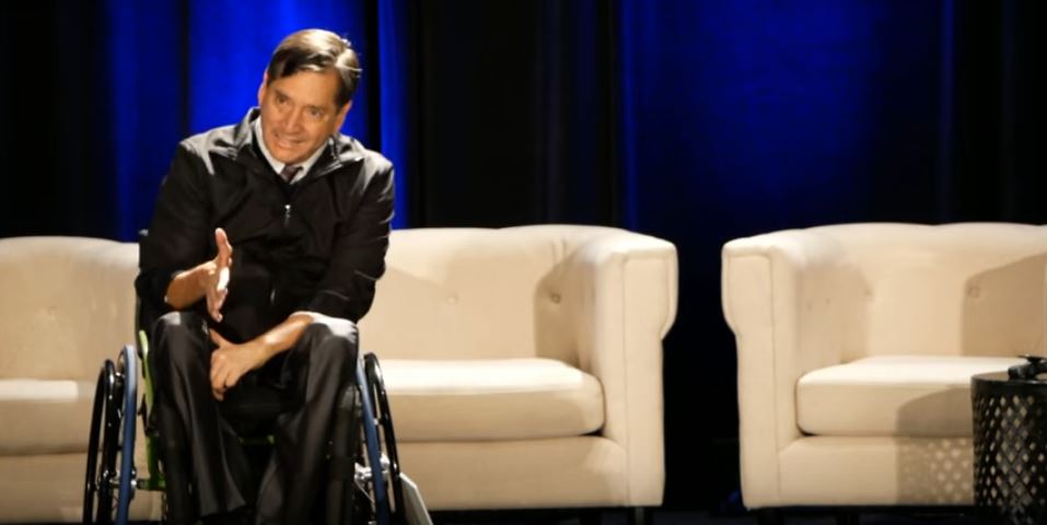 Santiago is seated in a wheelchair, motioning with his hands as he speaks. He is on a stage with couches behind him.