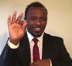 Close up of Buli, a black man in his 30s perhaps who is signing with his hand, wearing suit and tie