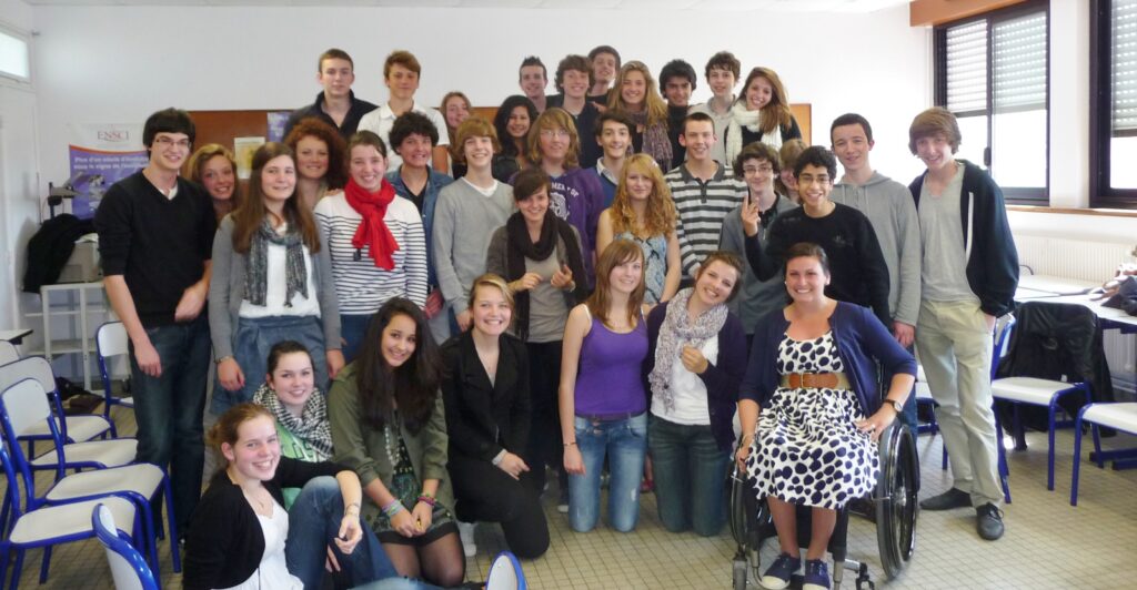 A young white woman in a manual wheelchair sits before a large group of perhaps 30 teens in a classroom