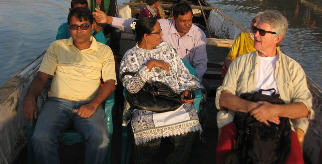 A small group of people are seated in a boat on a river. Most are people of color wearing dress common to Bangladesh. A white man with grey hair sits to the far right.