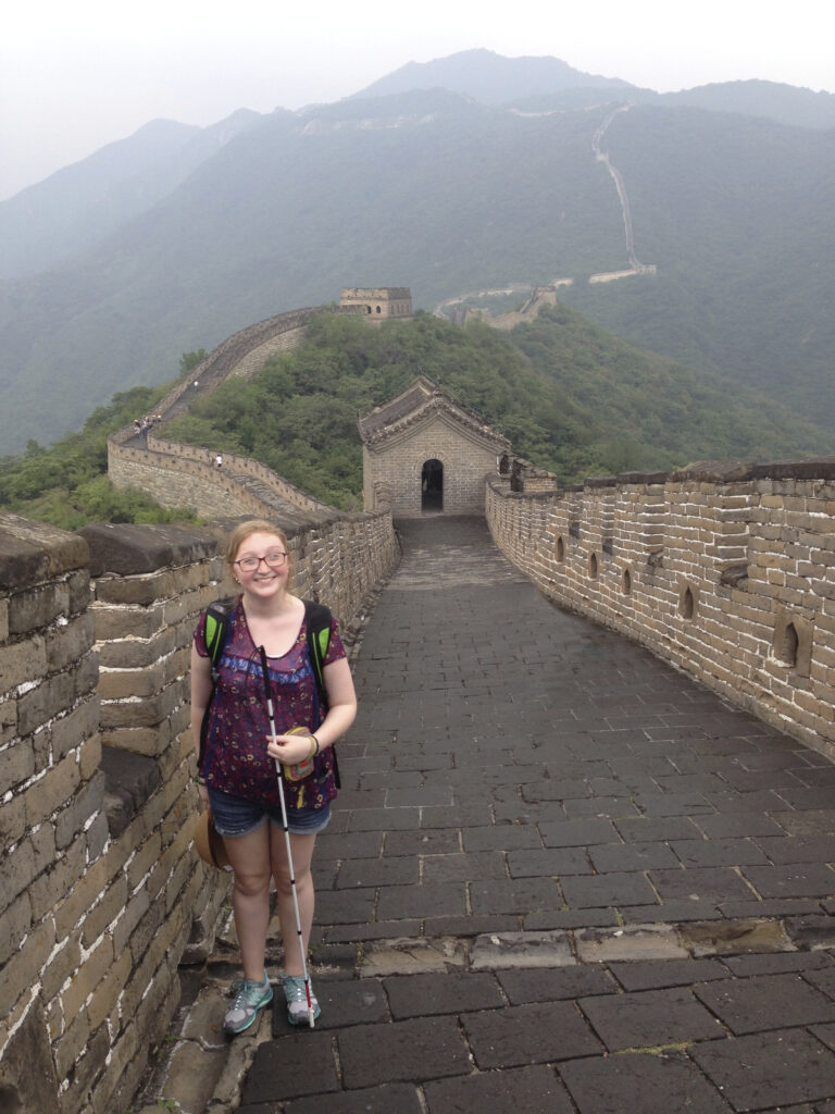 A young white woman stands with a white cane atop the Great Wall of China