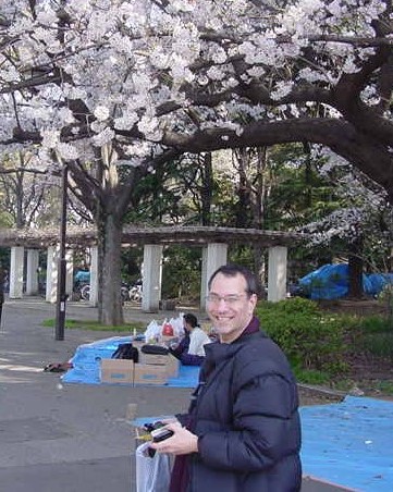 Kenny, a white middle aged man, looks over his shoulder smiling while beneath cherry blossom tree wearing winter coat