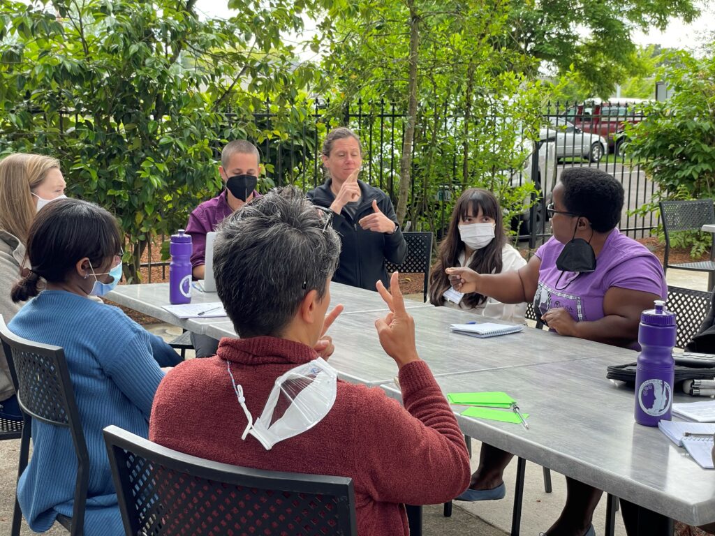A group of women seated at a table, some wearing covid masks, some signing in sign language. 