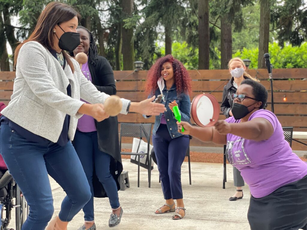A group of women dance together, shaking or beating musical instruments. Some women wear purple WILD logo t-shirts, while others wear business casual clothing. Some women are non-disabled women, others are women with non-apparent disabilities, and another woman is short stature.