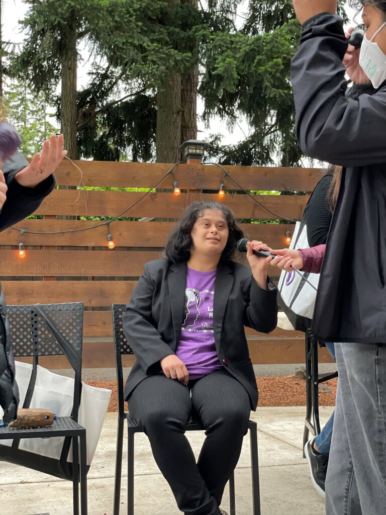 A woman with Down syndrome wearing a WILD logo t-shirt under a blazer is seated and speaks into a microphone on an outdoor patio.