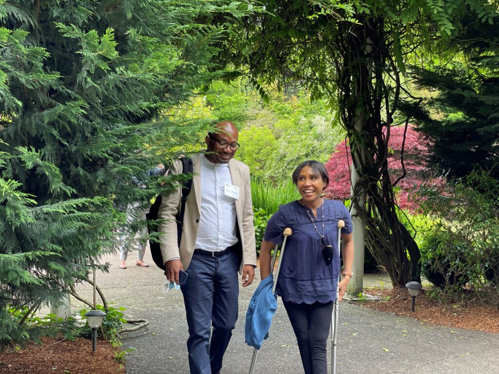 A man and a woman walk together along a garden path, smiling as they talk. The woman walks with crutches.