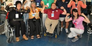 Four people including three women and three wheelchair riders pose for a photo in a fitness room flexing their forearms and smiling