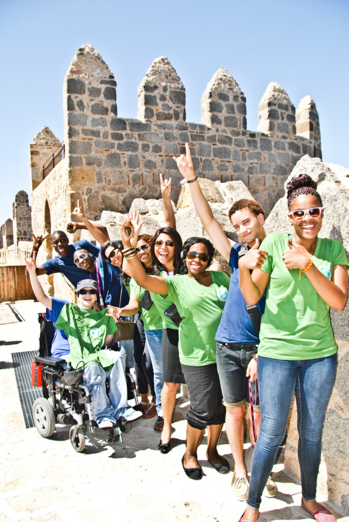 A group of ten or so young people in front of a castle on a clear day smile and wave energetically at camera. Many are people of color and one person uses a power wheelchair while others stand.