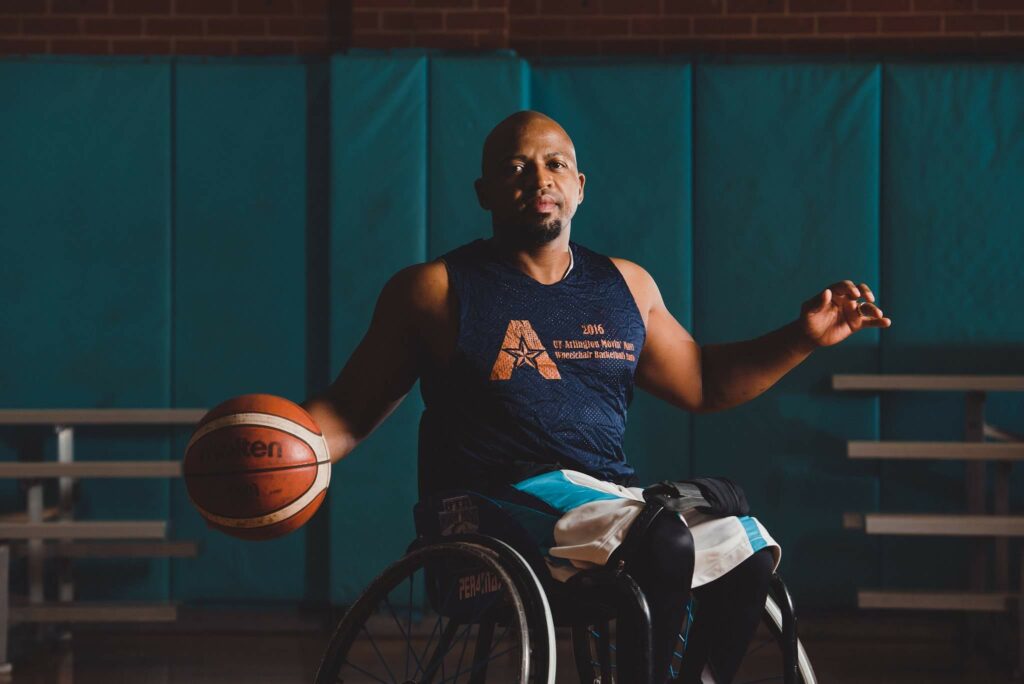 A portrait of Sips in a navy blue basketball uniform, a basketball wheelchair. He has his hands outstretched to the sides and holds a basketball in one. Behind him is a dark teal soft wall.