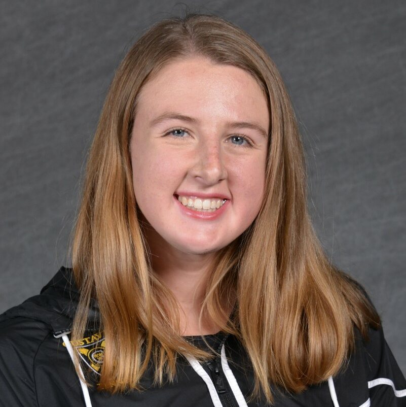 Hannah is wearing a black windbreaker from her college tennis team and is smiling for her yearly team photo.