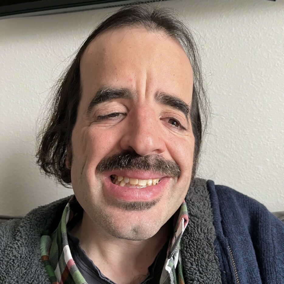 Close up of Trevor, his hair is straight and he wears a blue sweater.