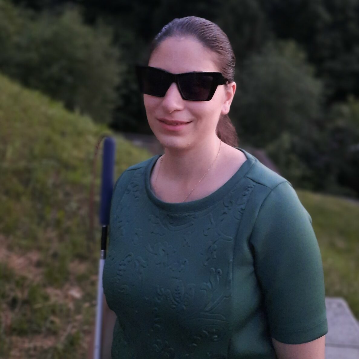 Portrait of Mariam behind her green forest. She had a pony tail, dark glasses, she wears a long sleeve dark green shirt and holds a white cane.
