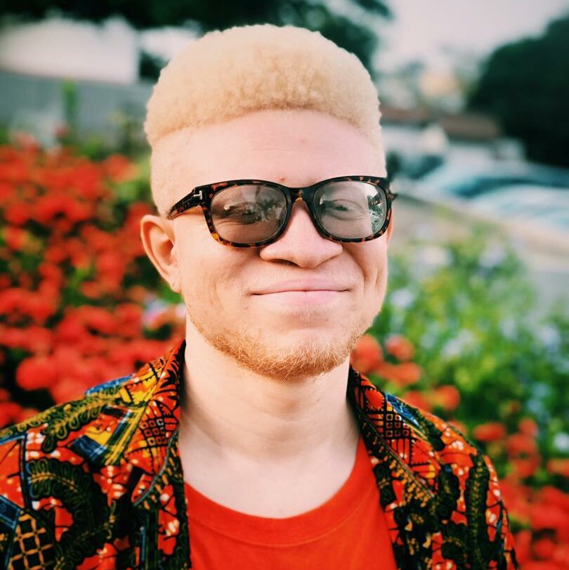 A portrait of Prince-Obed smiling at the camera. He has blonde hair and wears an abstract art shirt with an orange tank top under behind him orange flowers.
