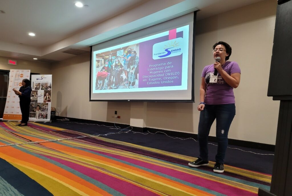 WILD-El Salvador leader Valentina holds a microphone while presenting in a large room. Behind her to her right is a projector screen showing a photo of WILD in Eugene.