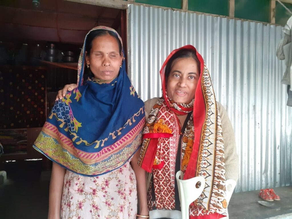 Two women stand together in front of a building wearing headscarves. One of those women is Sharmin, leader of WILD-Bangladesh. Sharmin has her arm around the shoulders of the other woman, a participant of WILD-Bangladesh.